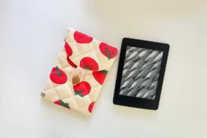Finished handmade nubim Kindle sleeve with tomato print fabric and wooden button loop closure placed beside a Kindle Paperwhite on a white surface