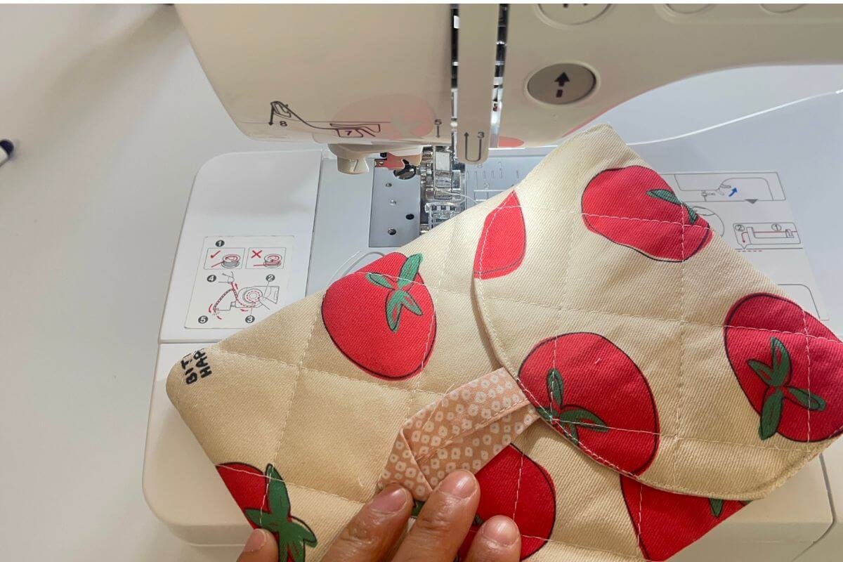 Hands guiding the nubim Kindle sleeve through a sewing machine while topstitching around the rounded flap edge, with the pink floral lining loop visible