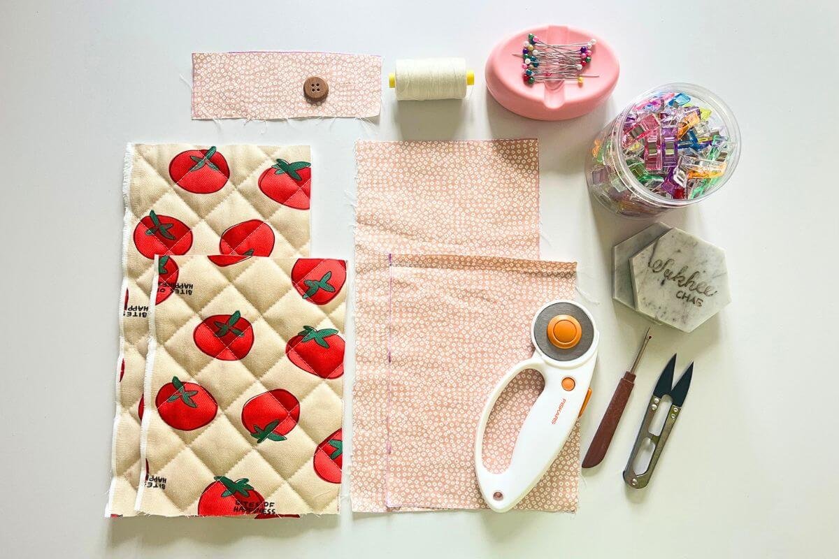 Flat lay of all sewing supplies needed for a nubim Kindle sleeve including cut nubim pieces, pink floral lining fabric, button, white thread, rotary cutter, seam ripper, pins and sewing clips on a white surface