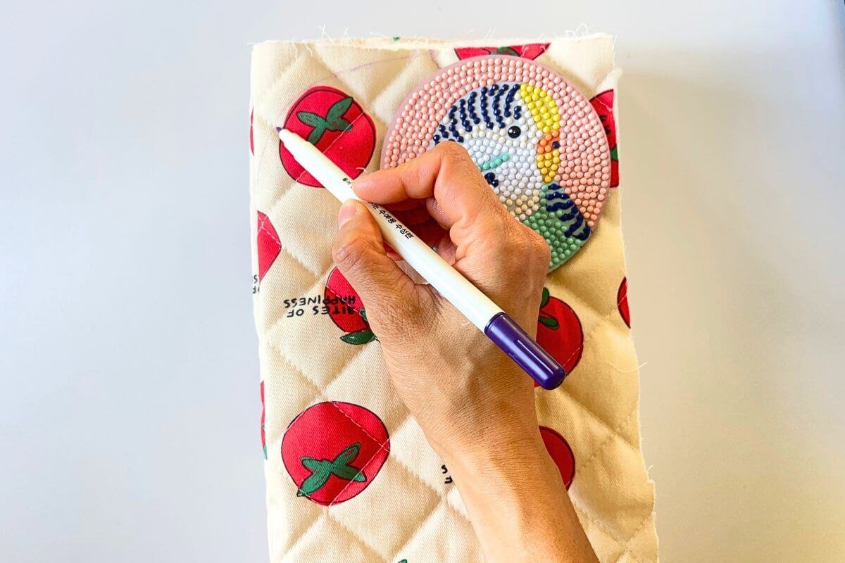 Hand holding a fabric marker tracing around a round decorative coaster to mark a curved corner on tomato print nubim quilted fabric